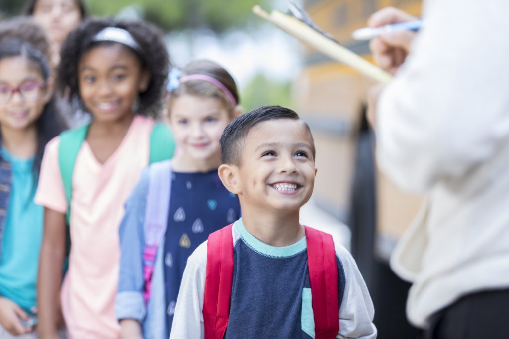 Unrecognizable school bus driver holds a clipboard and checks off each child as they get on a school bus. The children are lined up waiting to load the bus.