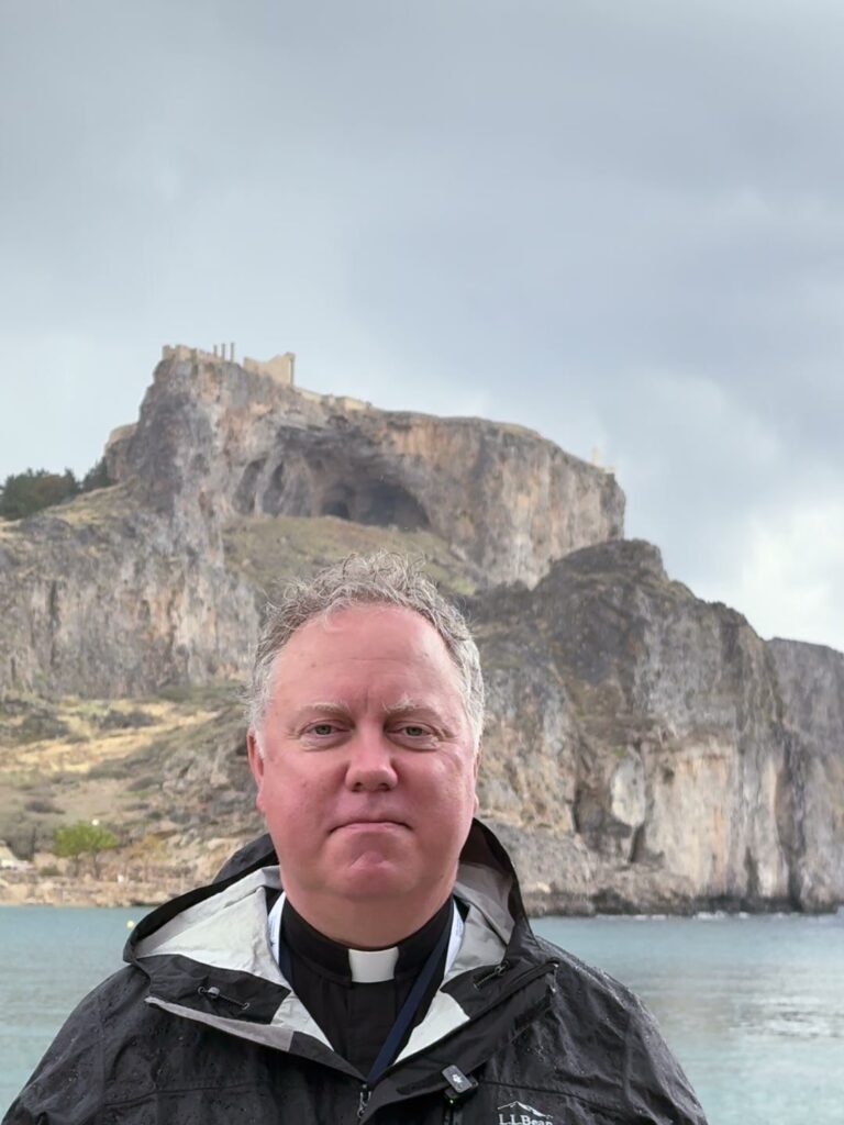 Fr. Jonathan Wilson at St. Paul’s Bay with the acropolis of Lindos behind him