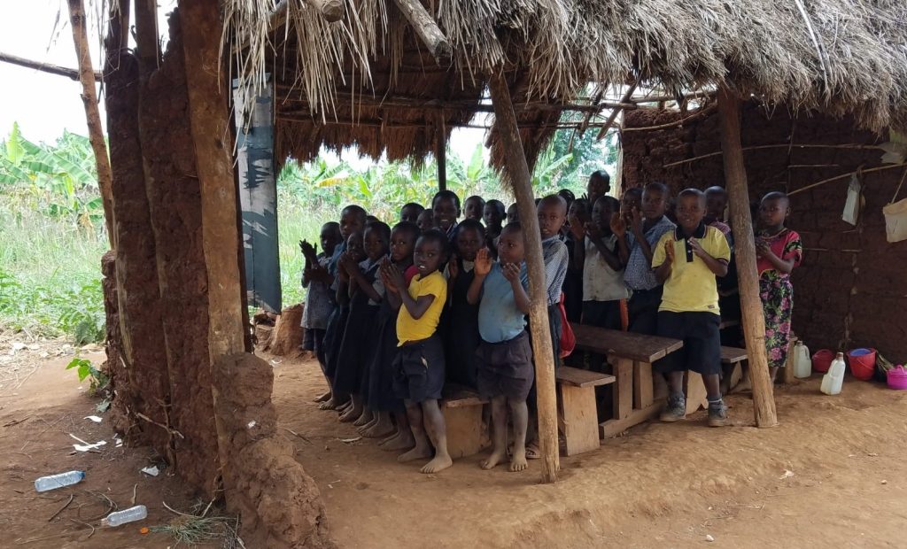 students stand under grass roofed cover with only 2 walls