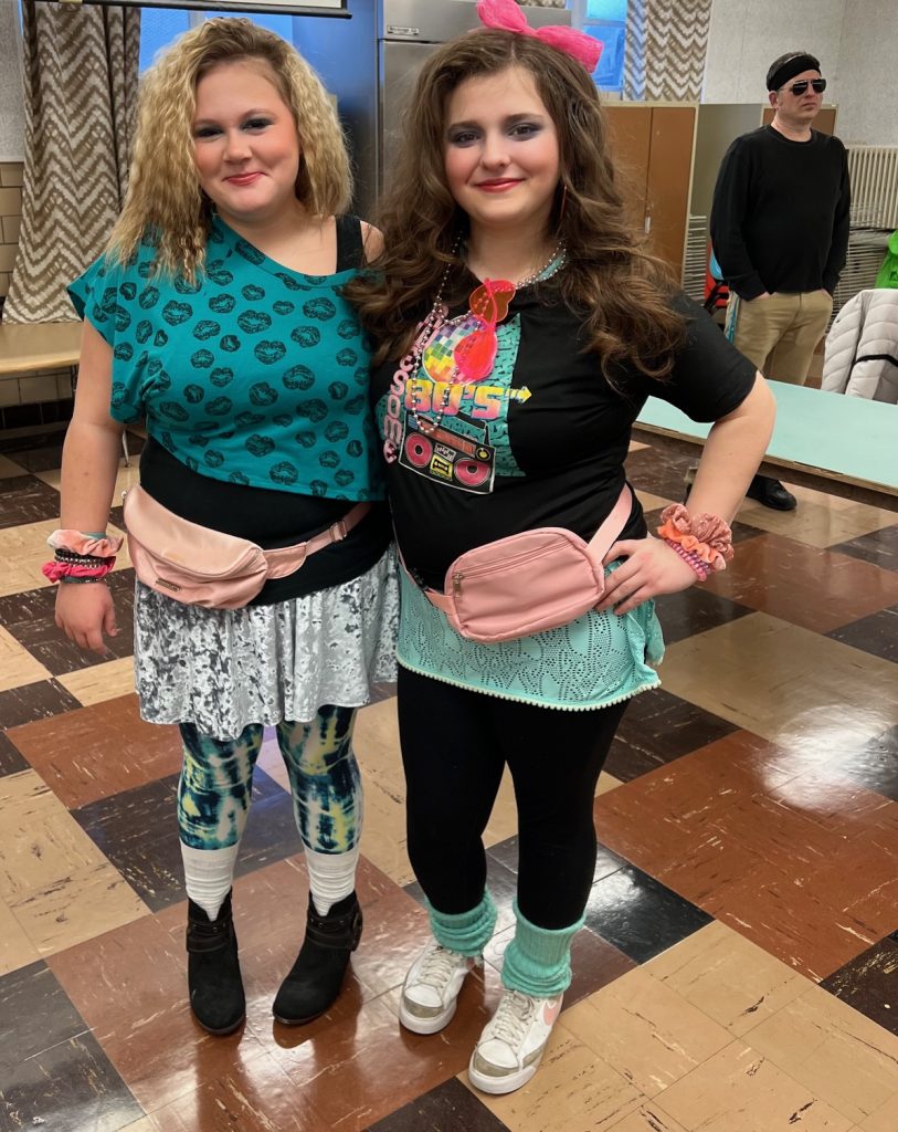8th grade students dressed in 80s apparel, posing smiling in the school cafeteria
